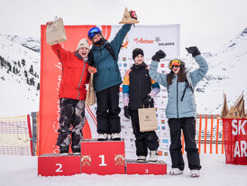 Vier Mädchen feiern ihren Erfolg in der Kategorie U14 mit hochgestreckten Armen auf dem Siegerehrungspodest. Im Hintergrund ist die verschneite Berglandschaft sichtbar.