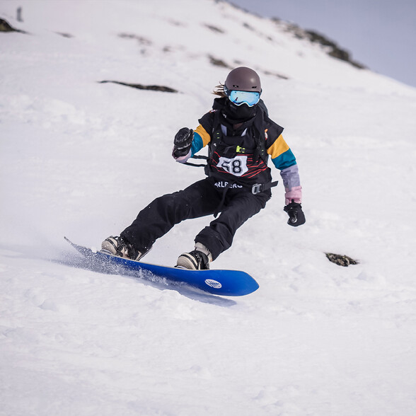 Eine Snowboarderin fährt dynamisch eine verschneite Abfahrt hinunter. Sie trägt einen Helm, eine Schutzbrille und eine mehrfarbige Snowboardjacke.
