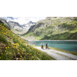 Zwei Radfahrer auf Schotterweg entlang eines blauen Stausees inmitten grüner Berglandschaft / Two cyclists on gravel path along a blue reservoir surrounded by green mountain scenery