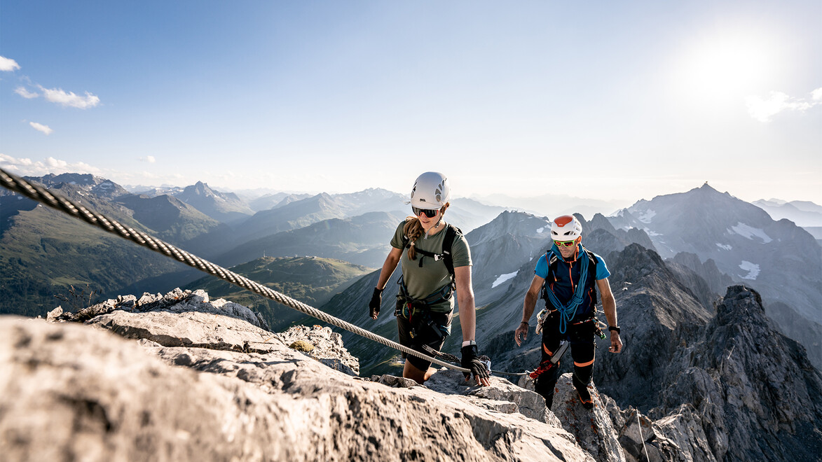 Zwei Klettersteiggeher mit Helm und Ausrüstung auf luftiger Felspassage mit Panoramablick / Two climbers with helmet and gear on exposed rocky section with panoramic view
