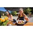 Zwei Frauen essen unter Sonnenschirm auf Holzbank bei Hütte, eine serviert Salat / Two women eat under parasol on wooden bench at a hut, one serving salad