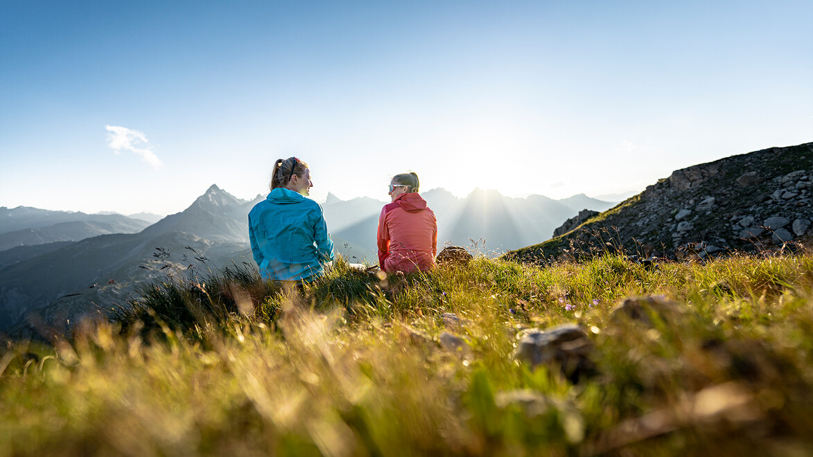 Zwei Personen sitzen auf blühender Almwiese und blicken in die Ferne auf das Abendlicht / Two people sit on a flowering alpine meadow gazing into the distance at the evening light