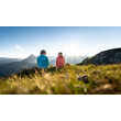 Zwei Personen sitzen auf blühender Almwiese und blicken in die Ferne auf das Abendlicht / Two people sit on a flowering alpine meadow gazing into the distance at the evening light