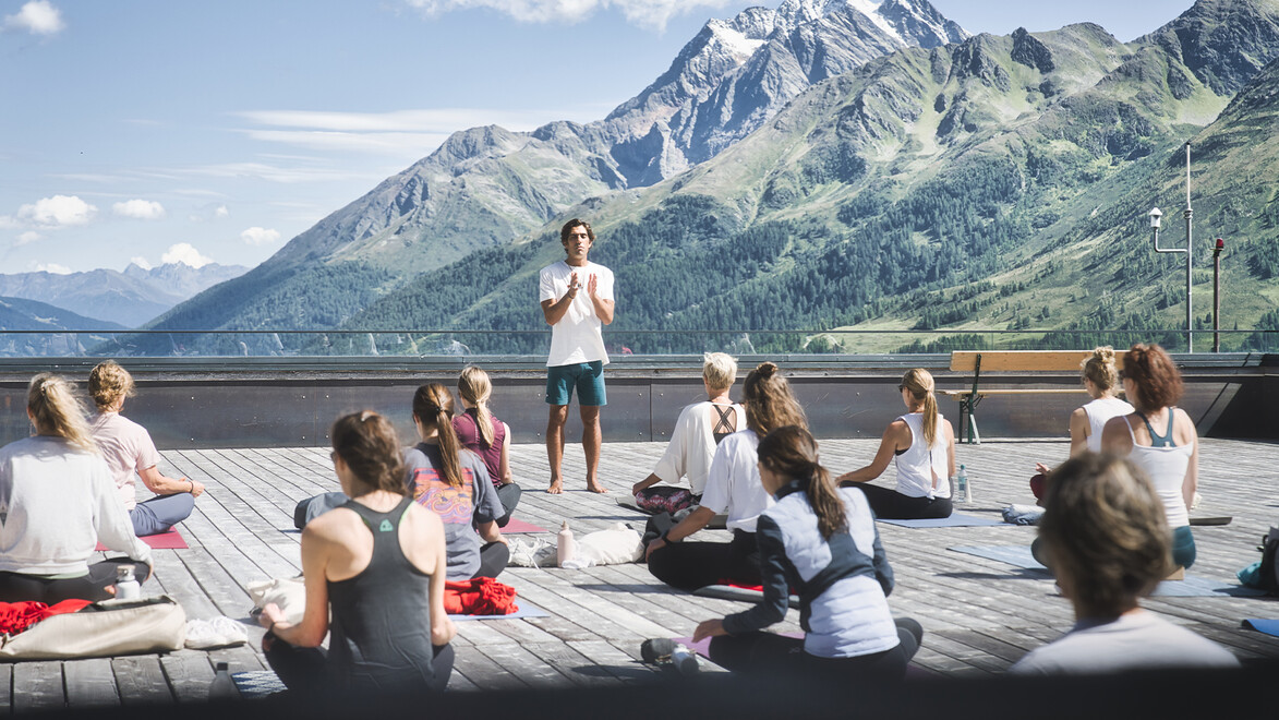 Gruppe bei Outdoor-Yoga auf einer Holzplattform vor Bergkulisse / Group during outdoor yoga on a wooden platform in front of mountain scenery