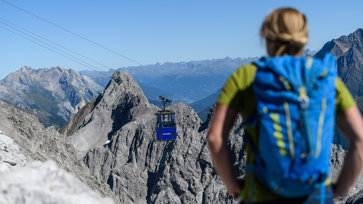 Eine Person mit Rucksack steht auf felsigem Terrain und blickt auf eine Seilbahn in alpiner Berglandschaft / A person with a backpack stands on rocky terrain and looks at a cable car in the alpine mountain scenery