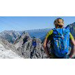 Eine Person mit Rucksack steht auf felsigem Terrain und blickt auf eine Seilbahn in alpiner Berglandschaft / A person with a backpack stands on rocky terrain and looks at a cable car in the alpine mountain scenery