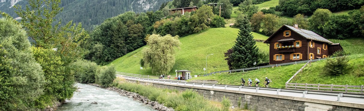 Gruppe von Radfahrern fährt auf einer einem Weg ad bei Flirsch neben der der Rosanne  t durch graue en h hügel mit Holzhäusern.