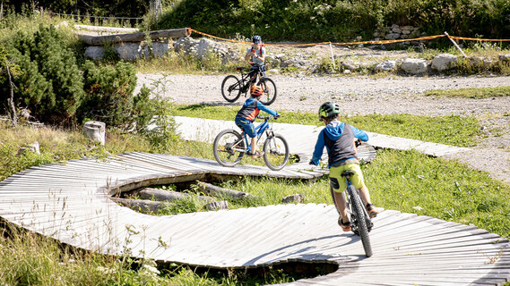 Drei Kinder fahren mit dem Mountainbike auf einem geschwungenen Holzweg im grasbewachsenen EldoRADo Bike-Areal Verwall