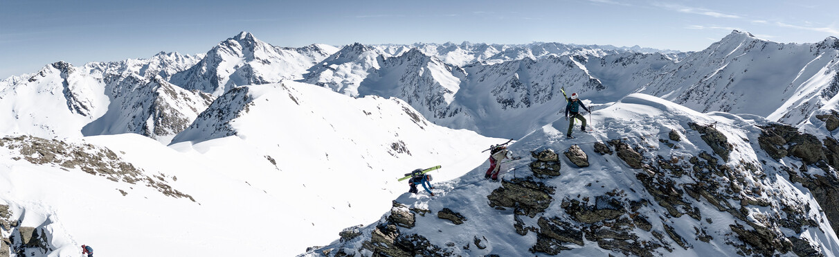 Three freeriders secure themselves on a snowy ridge with panoramic views of the surrounding Alpine peaks.