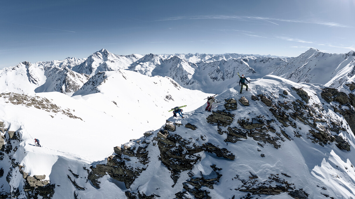 Three freeriders secure themselves on a snowy ridge with panoramic views of the surrounding Alpine peaks.