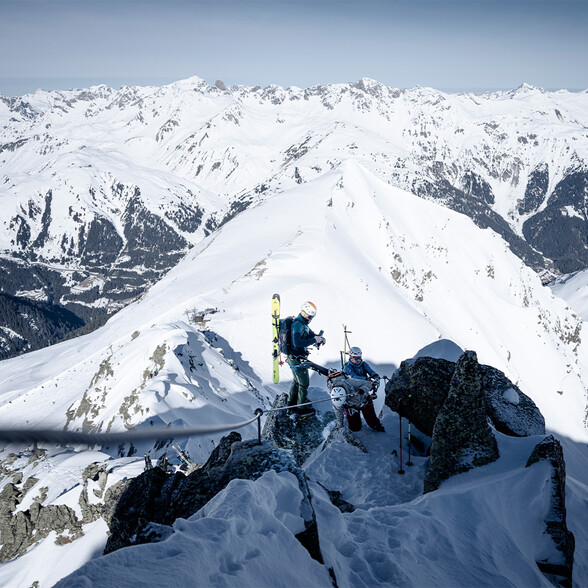 Two freeriders on a summit slope with stunning views of the snowy Stanzertal valley and surrounding peaks.