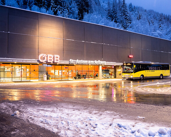 Der beleuchtete Vorplatz des Bahnhofs St. Anton am Arlberg bei Dämmerung mit ÖBB-Schriftzug, gelbem Linienbus und spiegelndem Schnee auf dem Boden.