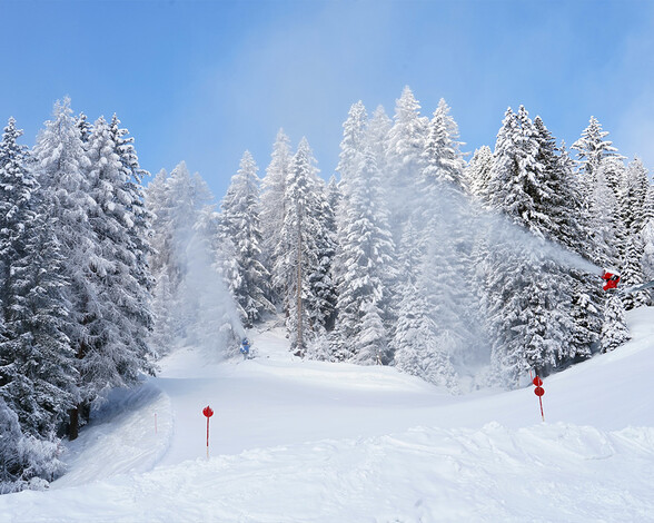 Eine aktive Schneekanone beschneit eine winterliche Waldlichtung in St. Anton am Arlberg unter blauem Himmel und mit frisch verschneiten Bäumen.