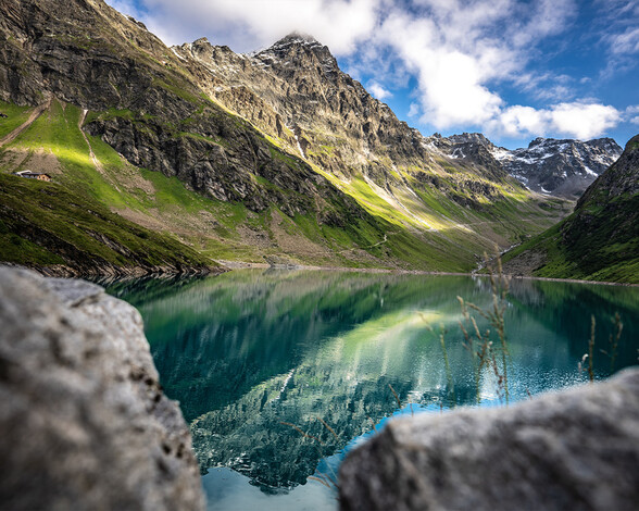 Der Kartellsee liegt eingebettet in grünen Berghängen unter blauem Himmel. Das klare Wasser spiegelt die umliegenden Berge eindrucksvoll wider.