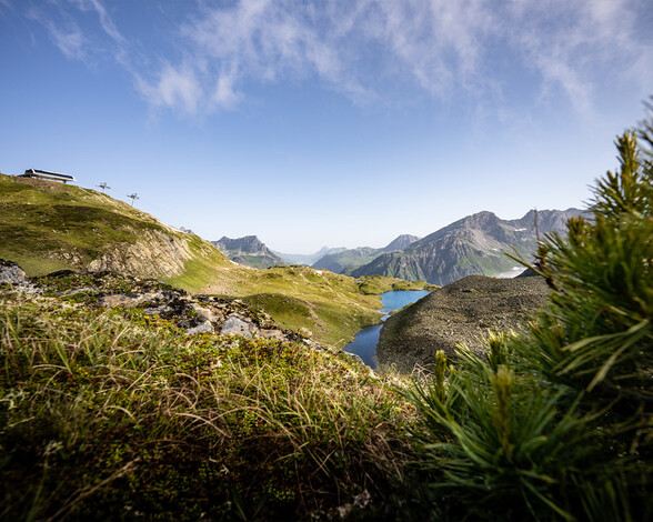 Hoch über St. Anton am Arlberg zeigt das Bild ein grünes, alpenblumenreiches Sommerpanorama mit Blick auf Bergseen und die Liftstation.
