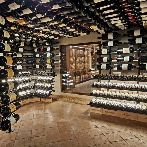 Ein luxuriöser Weinkeller mit deckenhohen Weinregalen, in denen zahlreiche Weinflaschen gelagert sind. Die Flaschen liegen horizontal und sind optimal beschriftet. Durch ein großes Fenster ist ein angrenzender Raum mit weiteren Weinregalen sichtbar.