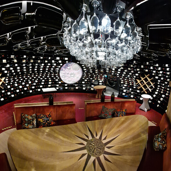 Ein eindrucksvoller Weinkeller mit hunderten Weinflaschen, einem modernen Kronleuchter aus Gläsern und einem runden Sitzbereich mit rotem Teppich. Im Zentrum steht ein Tisch mit Windrosen-Intarsie. Der Raum ist elegant gestaltet und ideal für exklusive Weinverkostungen.
