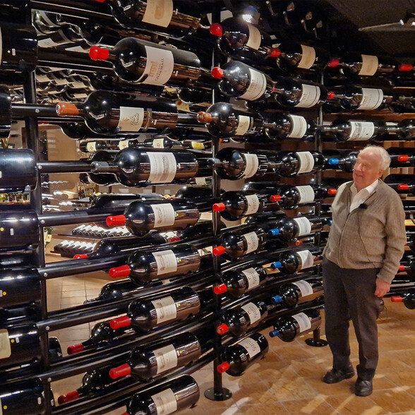 Ein älterer Mann steht in einem exklusiven Weinkeller vor einer Wand mit zahlreichen, horizontal gelagerten Weinflaschen. Die Flaschen sind ordentlich sortiert und spiegeln sich im glänzenden Regal. Der Raum ist stimmungsvoll beleuchtet und vermittelt ein Gefühl von Eleganz und Weinkultur.