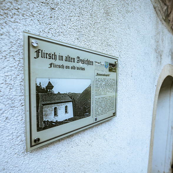 Eine Infotafel in Flirsch an der Außenmauer einer Kapelle zeigt historische Einblicke in die Antoniuskapelle mit Bild und Text.