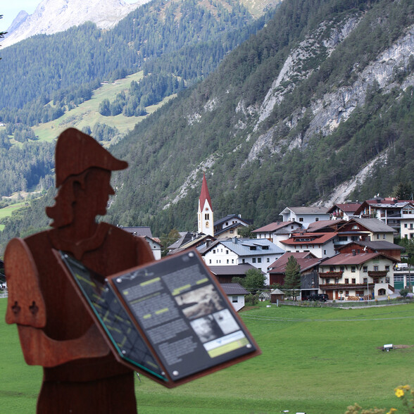Eine Silhouette aus Metall mit Infotafel steht am Schnanner Themenweg mit Blick auf das Dorf Schnann und die Berge im Hintergrund.