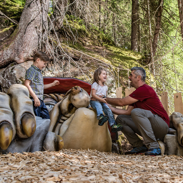 Ein Vater spielt mit zwei kleinen Kindern auf einer fantasievollen Spielstation im Wald. Die Kinder sitzen lachend auf einer großen, trollartigen Handfigur. Die Umgebung besteht aus Holzspänen, Baumstämmen und Wurzeln – ideal für ein Naturabenteuer.