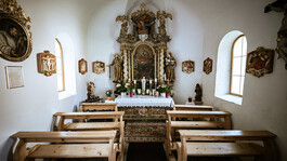 Blick in eine kleine Kapelle mit geschnitztem Altar, Holzbankreihen und religiösem Schmuck.