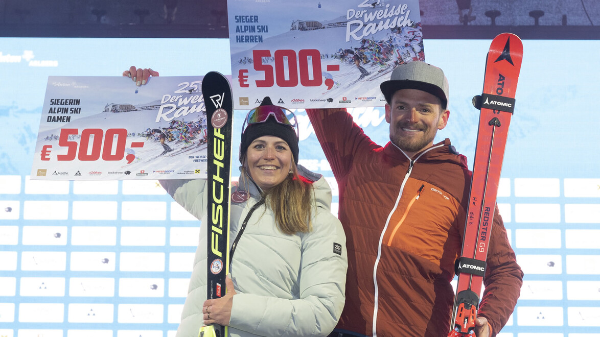 Frau und Mann mit Skiern und Gewinngutscheinen lächeln vor Siegerehrungstafel / Woman and man with skis and winner vouchers smiling in front of results board