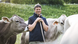 Ein Landwirt steht auf einer grünen Almwiese zwischen Kühen und blickt lächelnd in die Kamera.
