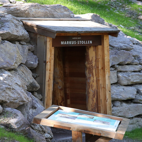 Ein kleiner Stolleneingang aus Holz mit der Aufschrift „Glück auf! Markus-Stollen“ ist in eine Felswand eingebettet. Davor steht eine hölzerne Infotafel über den historischen Bergbau.