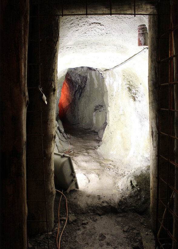 Blick in das Innere eines historischen Bergwerksstollens mit weißem Gestein. Die Gänge sind uneben und mit Stromleitungen ausgestattet, schwaches Licht erhellt den Weg.