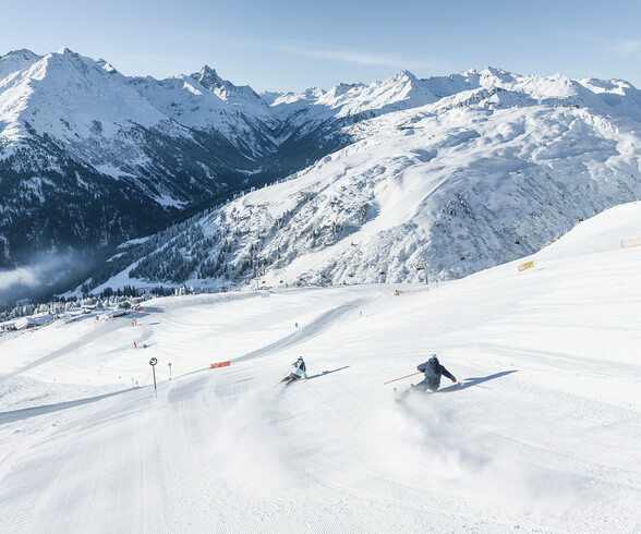 Zwei Skifahrer fahren bei Sonnenschein eine frisch präparierte Skipiste in St. Anton am Arlberg hinab. Im Hintergrund erstrecken sich die schneebedeckten Gipfel der Tiroler Alpen.