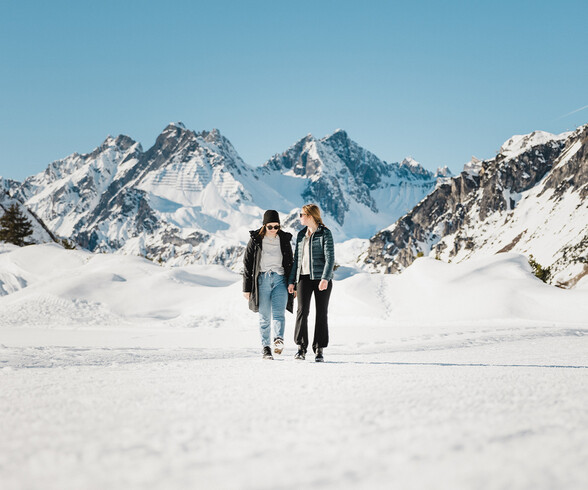 Zwei Frauen spazieren durch verschneite Landschaft mit imposanter Bergkulisse in St. Anton am Arlberg. Der blaue Himmel unterstreicht die klare Winterluft.