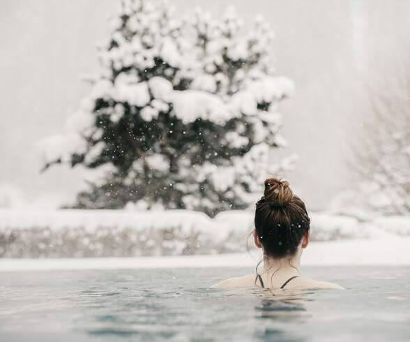 Eine Frau entspannt im beheizten Außenpool, während sanfte Schneeflocken fallen. Im Hintergrund steht ein schneebedeckter Nadelbaum – Wellness im Winter in St. Anton.