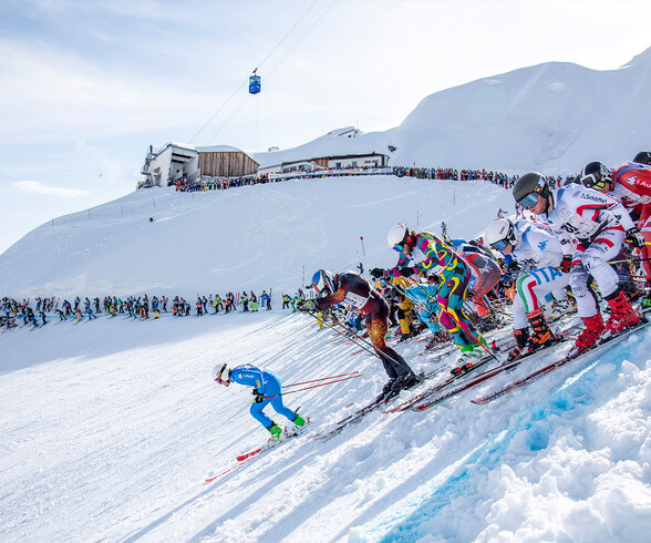 Zahlreiche Skifahrer starten gleichzeitig bei „Der Weiße Rausch“ in St. Anton. Die steile Piste und jubelnde Zuschauer im Hintergrund machen das Rennen zum Spektakel.