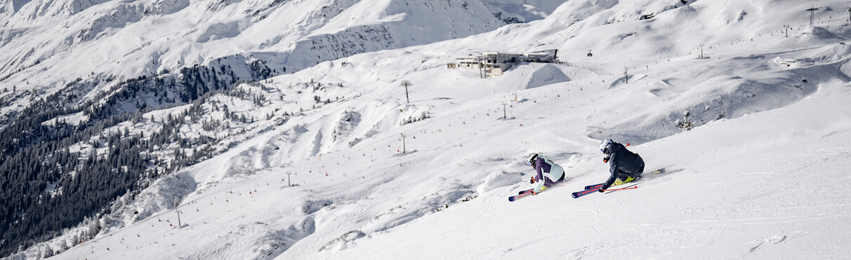 Zwei Skifahrerinnen carven parallel über eine breite, sonnige Piste mit Blick auf das winterliche Skigebiet in St. Anton.