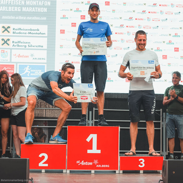 Die Sieger des Arlberg Marathons feiern auf dem Podest: Drei glückliche Läufer mit Urkunden und Preisen vor der Eventbühne.