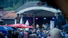 Besucher stehen vor einer Bühne, auf der eine Band bei einem Open-Air-Konzert auftritt / Visitors stand in front of a stage where a band performs at an open-air concert
