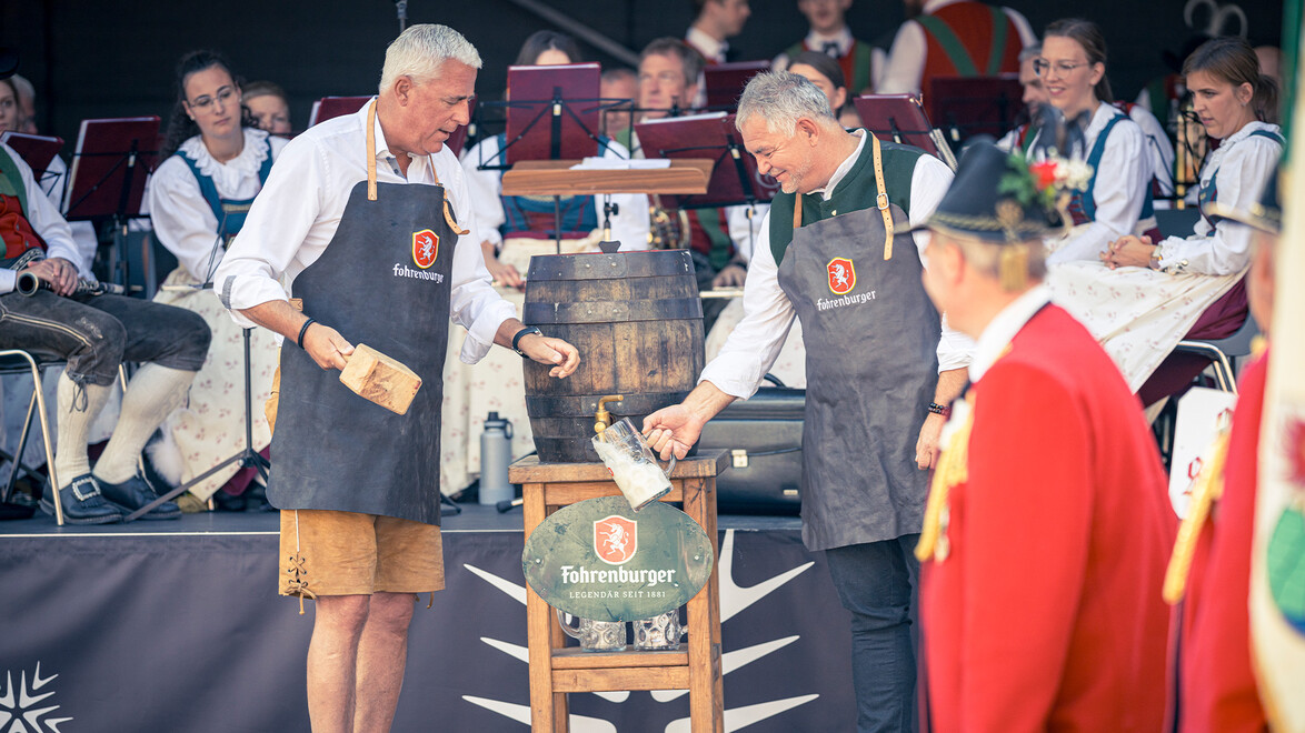 Zwei Männer zapfen gemeinsam ein Bierfass auf der Bühne eines traditionellen Festes an / Two men tap a beer keg together on the stage of a traditional celebration