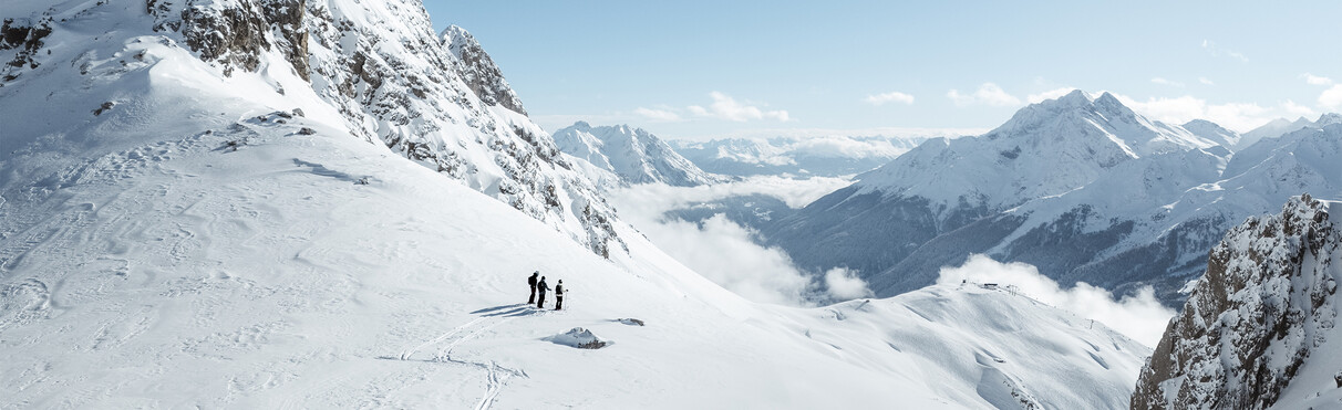 Drei Skifahrer stehen auf einem verschneiten Hochplateau mit Blick auf die Arlberger Bergwelt.
