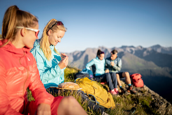 Four young hikers take a break on a sunny mountain summit in Tyrol. Two women in the foreground enjoy a snack, while the majestic Alps of St. Anton am Arlberg stretch out in the background – hiking with panoramic views in the Austrian mountains.