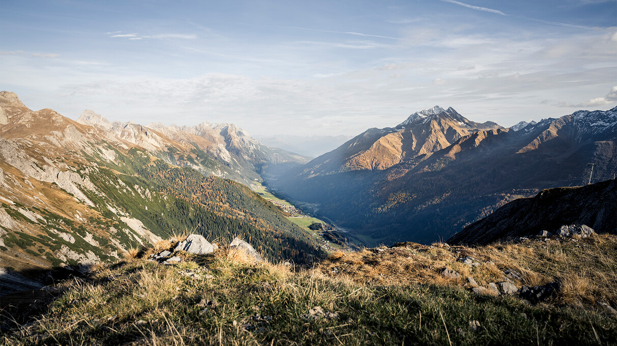 View into an autumnal Stanzertal valley with golden and green forests. The valley is surrounded by rugged mountains, some with light snow cover. The sky is bright with thin clouds.