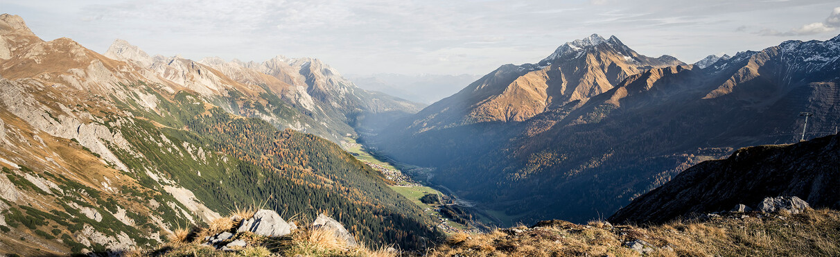Blick in das herbstliche Stanzertal mit goldgelben und grünen Wäldern. Umgeben ist das Tal von schroffen Bergen, einige mit leichtem Schneebedeck. Der Himmel ist hell mit dünnen Wolken.