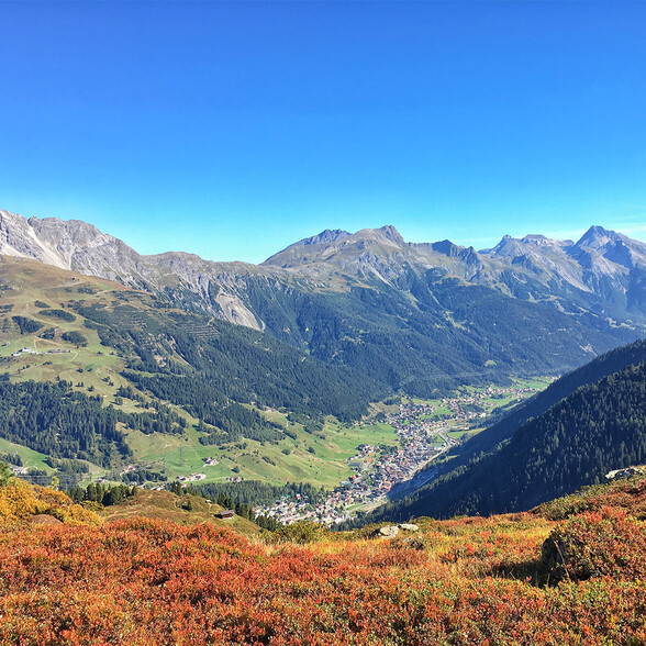 Herbstliche Berglandschaft mit roten Sträuchern im Vordergrund. Im Tal liegt ein Ort mit vielen Häusern. Dahinter erstrecken sich bewaldete Hänge und felsige Berggipfel unter blauem Himmel.