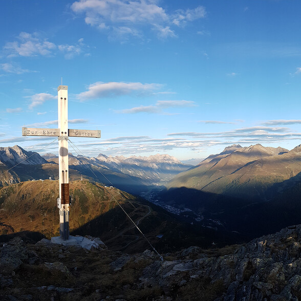 Ein weißes Gipfelkreuz steht auf einem felsigen Berggipfel. Im Hintergrund öffnet sich ein weites Tal mit vielen Bergketten. Der Himmel ist hellblau mit lockerer Bewölkung.