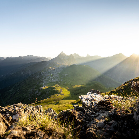 Sonnenstrahlen fallen über eine grüne, leicht felsige Berglandschaft. Im Hintergrund sind spitze Berggipfel zu sehen. Der Himmel ist hell, die Atmosphäre ruhig und klar.