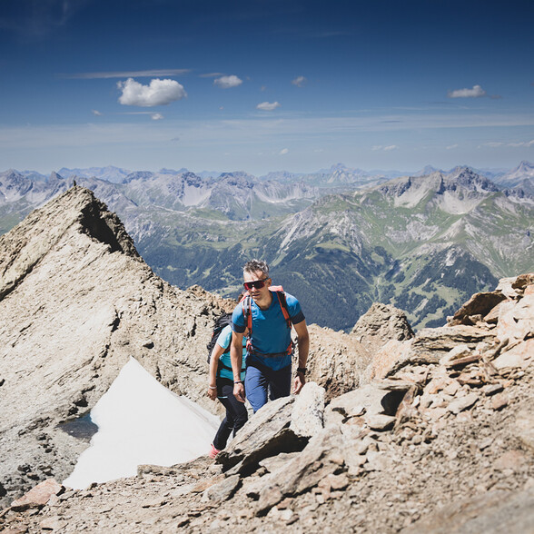 Zwei Bergsteiger in Funktionskleidung steigen über felsiges Gelände. Der Mann vorne trägt eine Sonnenbrille und einen roten Rucksack. Im Hintergrund sind steile Bergketten unter klarem Himmel sichtbar.