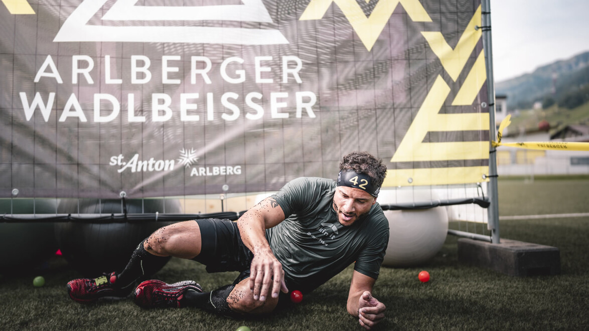 Ein Sportler kriecht lachend unter einem Netz mit dem Logo des Events im Hintergrund. / An athlete crawls laughing under a net with the event logo in the background.