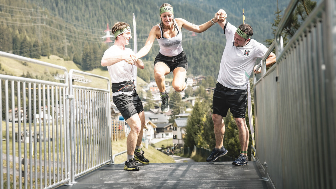 Zwei Personen helfen einer Teilnehmerin beim Absprung von einem Hindernis mit Panoramablick. / Two people assist a participant as she jumps off an obstacle with a panoramic view.
