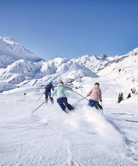 Skifahrer:innen gleiten bei Sonnenschein über eine perfekt präparierte Piste im Wintersportgebiet St. Anton am Arlberg.