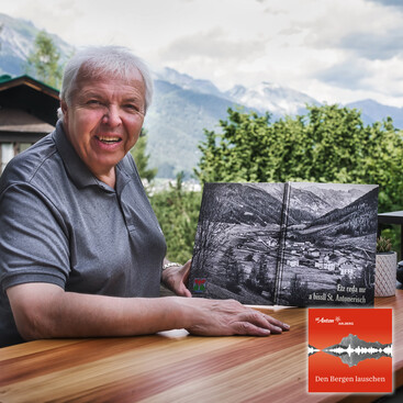 Andreas Fahrner sitzt an einem Holztisch im Freien mit Bergpanorama und hält ein großformatiges Buch mit dem Titel „Etz reda mr a bissl St. Antonerisch“, das eine schwarz-weiße Luftaufnahme von St. Anton zeigt.
